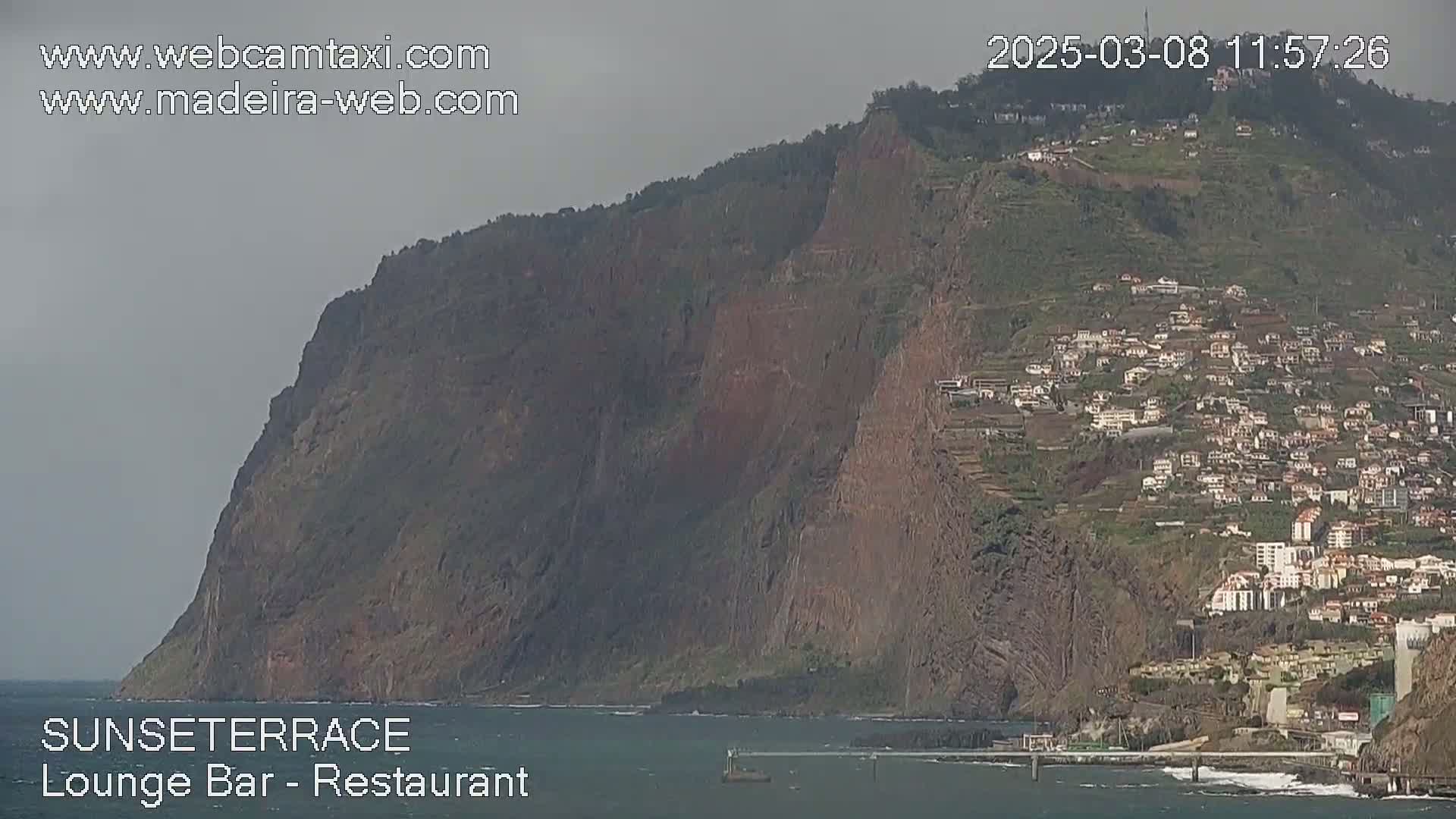 A large, dark cliff overlooks a town and the ocean under an overcast sky.