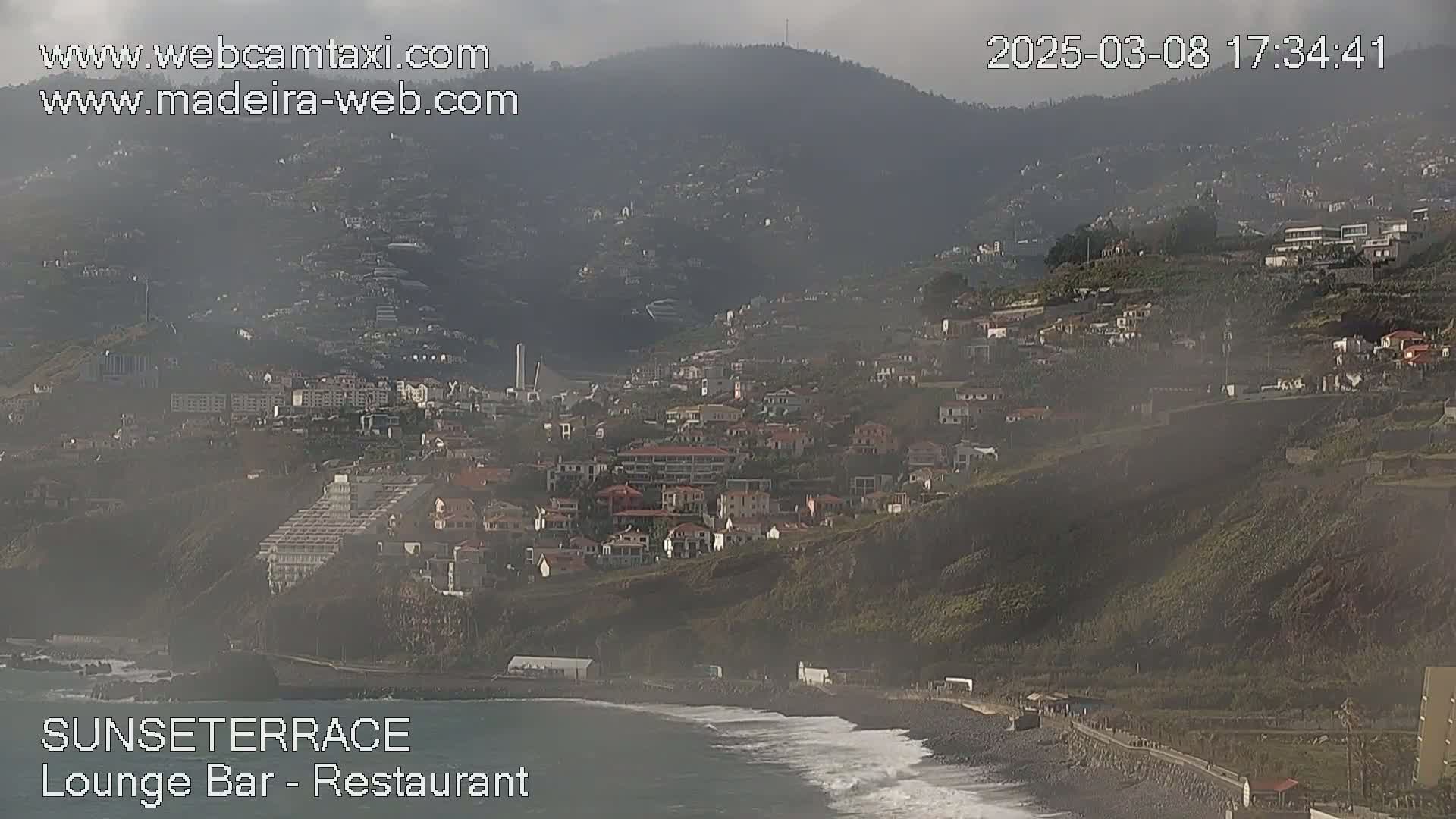 A coastal town nestled on a hillside with some fog is visible, overlooking a dark-sand beach with waves.