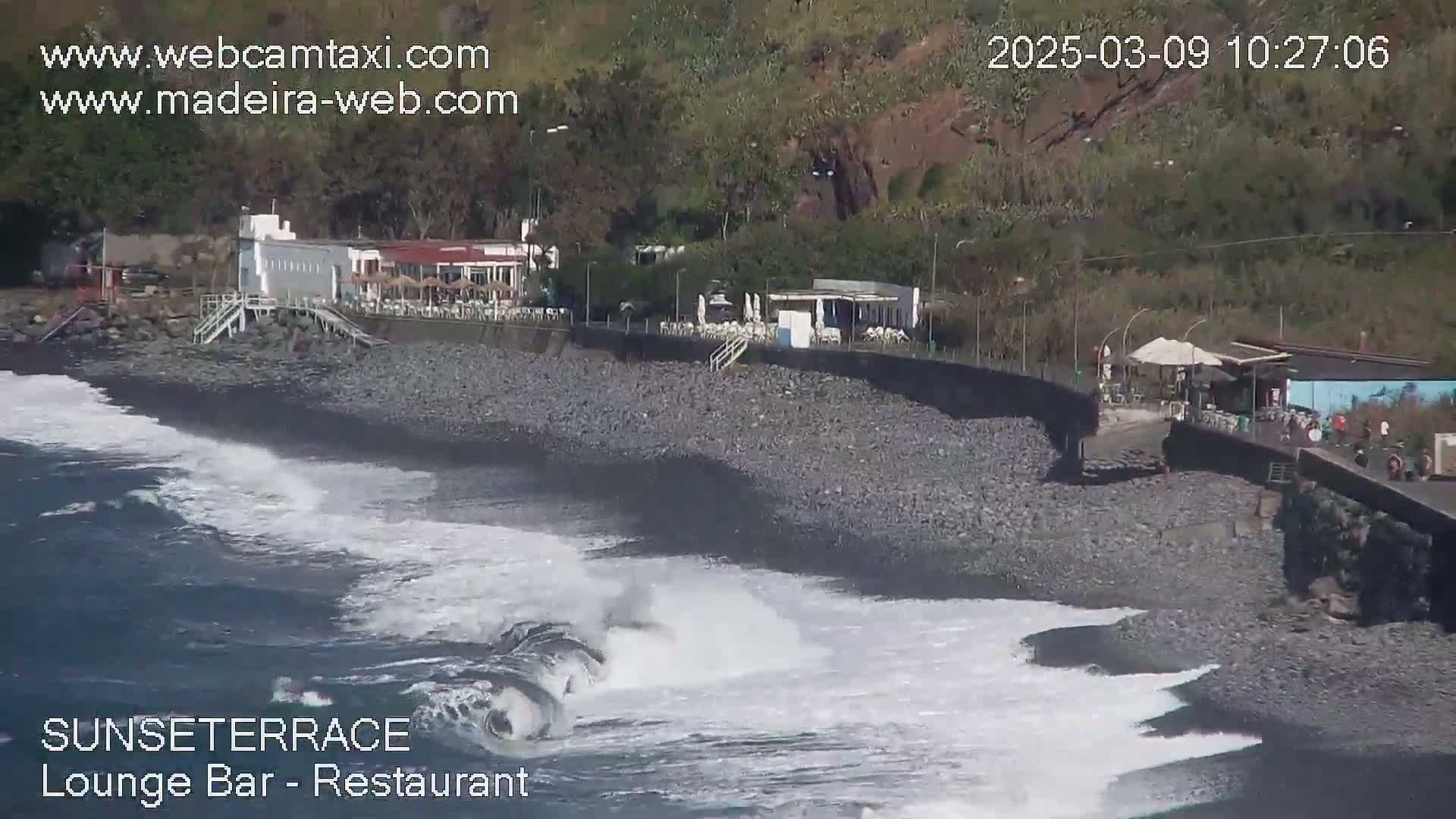 Ocean waves crash on a dark pebble beach near a shoreline restaurant and bar under a sunny sky.