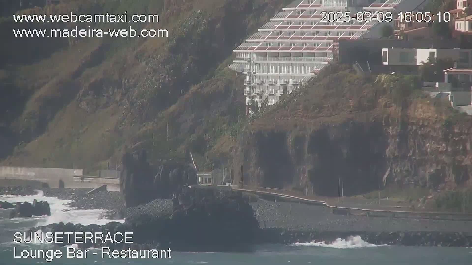 A multi-story white building sits atop a dark, rocky cliff overlooking a dark, pebble beach with small waves crashing against the shore.