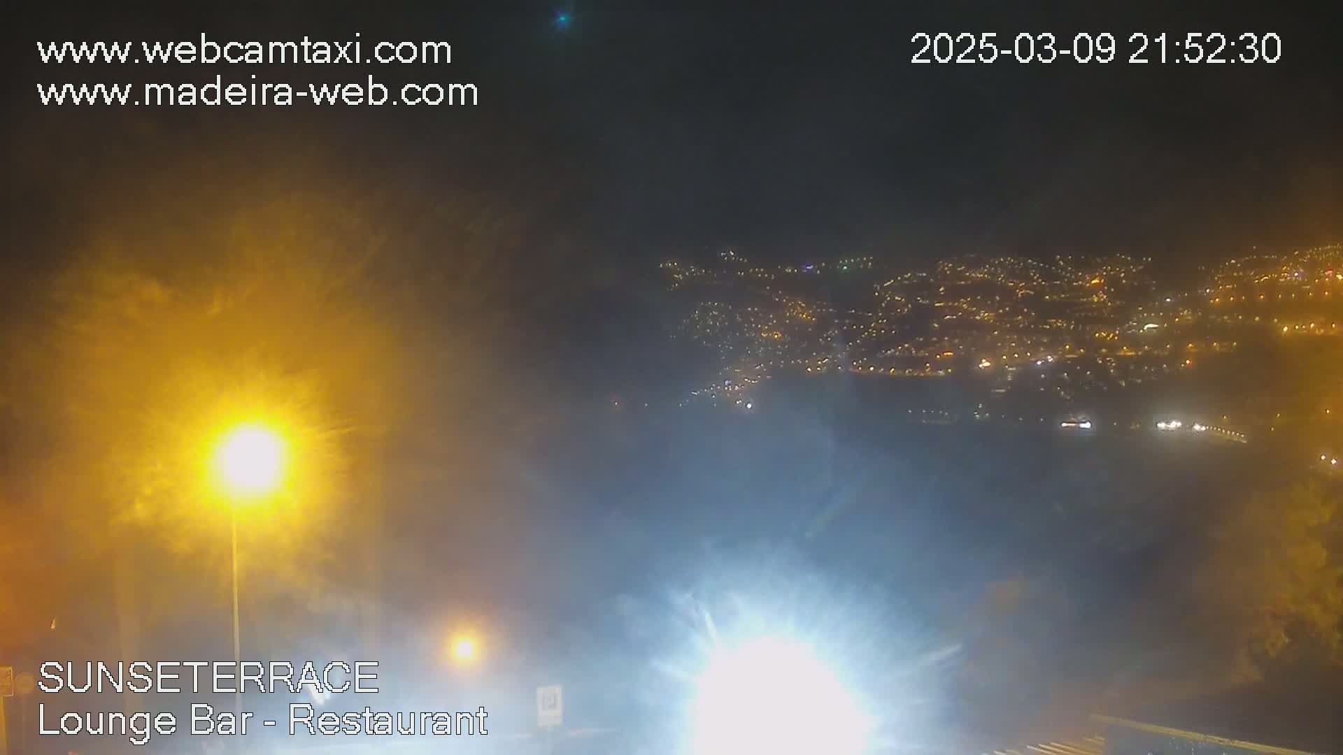 A hazy night scene shows a distant city illuminated with lights below, while a bright lamp post and some fog are visible in the foreground.