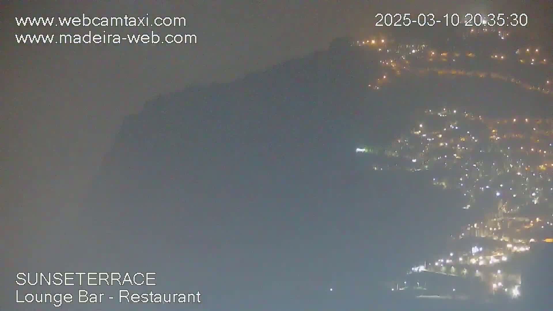 A hazy night scene shows a hillside town illuminated by numerous lights below a dark, hazy mountain.