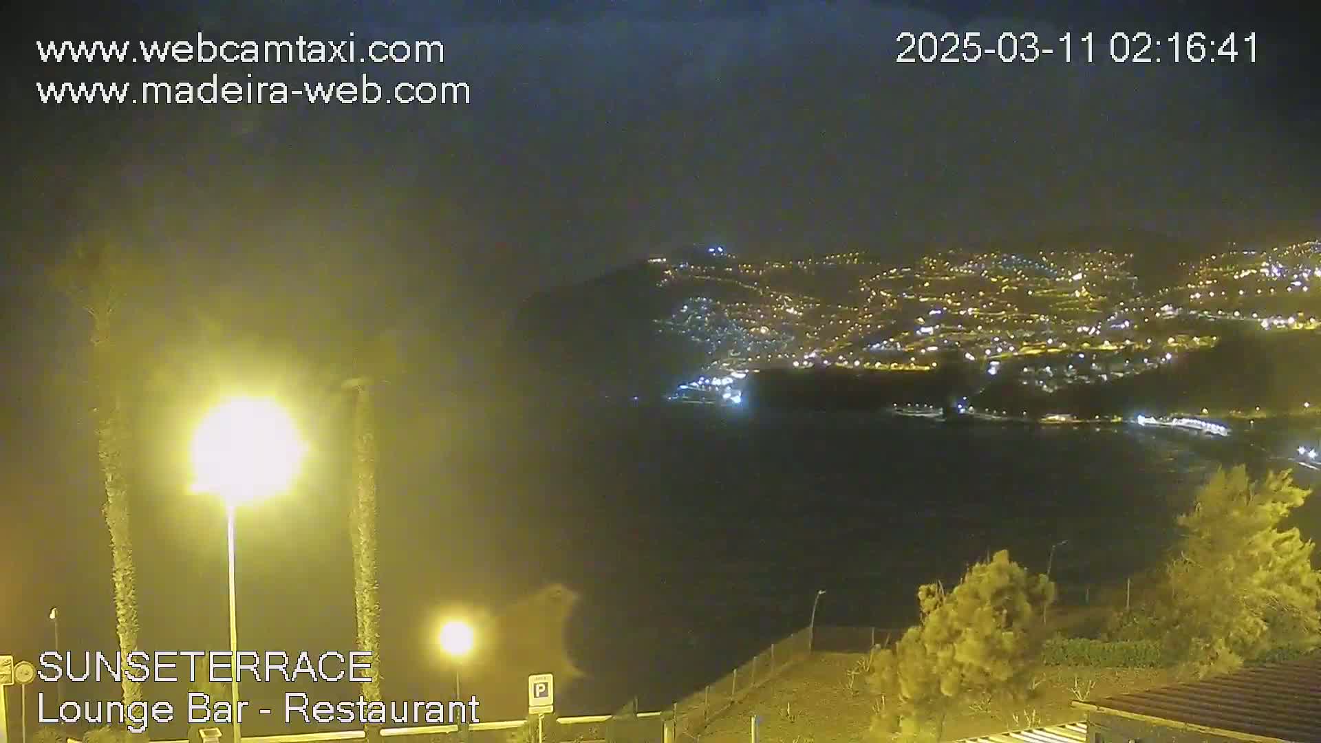 A nighttime, hazy view shows a brightly lit town nestled along a dark coastline, observed from a vantage point above.