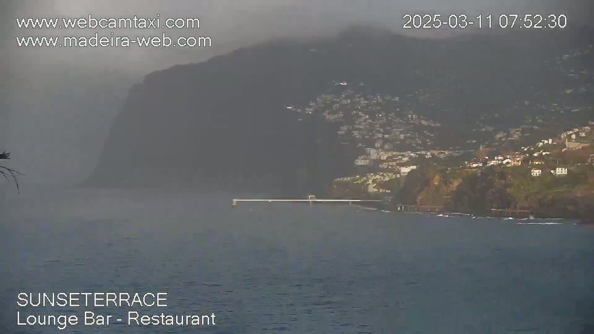 A hazy view of a coastal town nestled on a hillside overlooking a calm body of water with a long, thin pier extending into it.