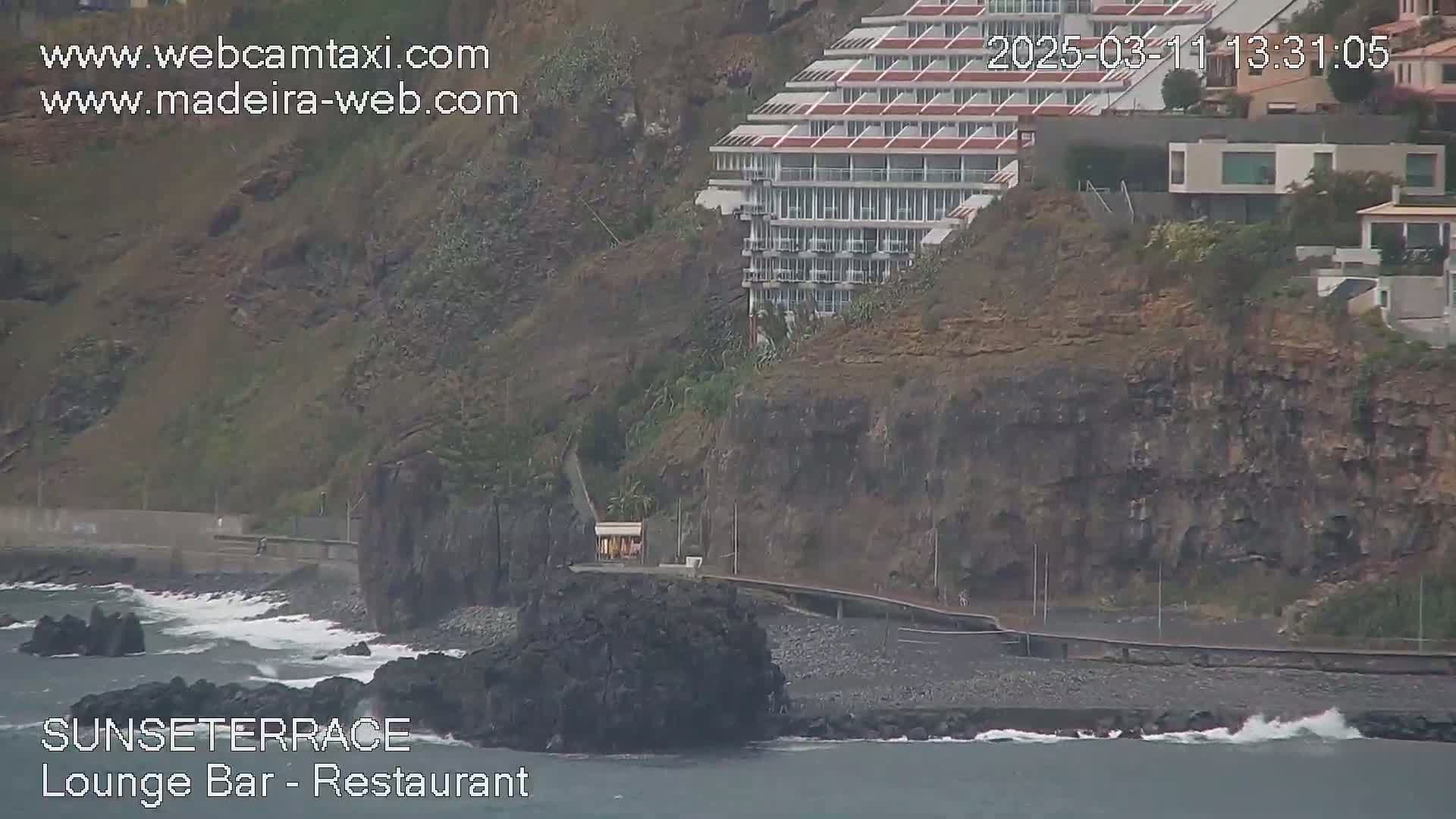 A multi-story building sits atop a dark cliff overlooking a rocky coastline with small waves crashing on the shore.