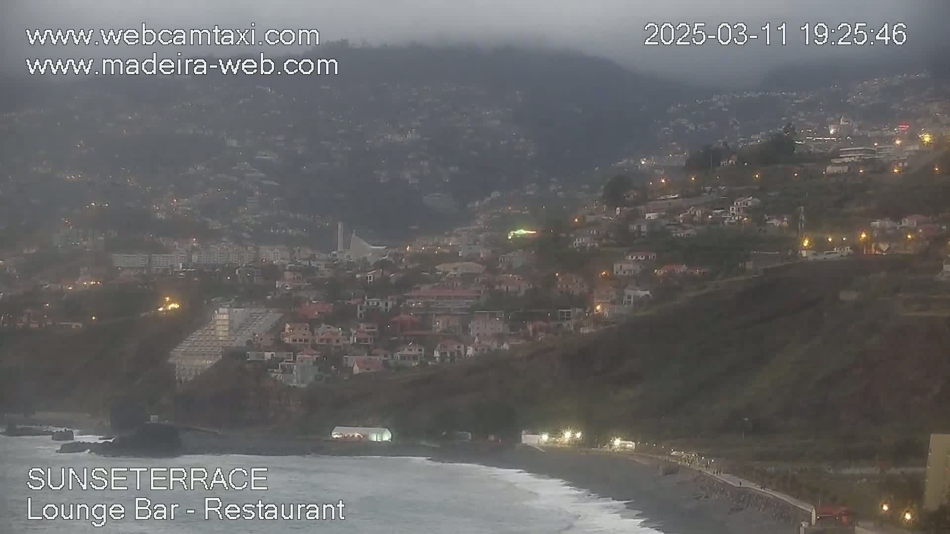 A coastal town at dusk is nestled on a hillside overlooking a dark beach with gentle waves under an overcast sky.