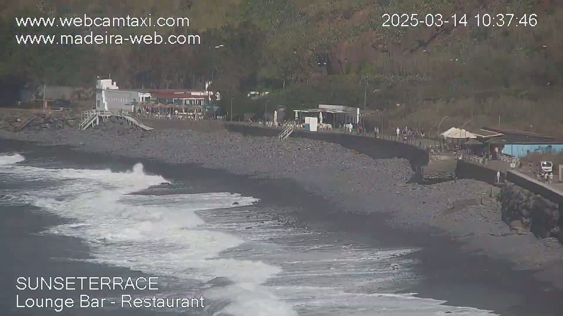 A dark-sand beach with small waves and a restaurant and bar next to a rocky hillside.