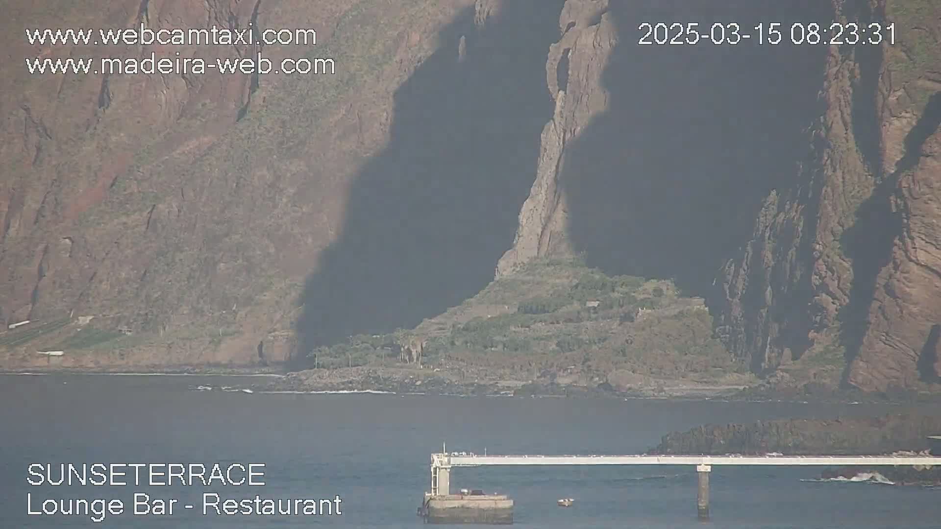 A long pier extends over calm water towards a rocky, vegetation-covered shoreline beneath a partly sunny sky.