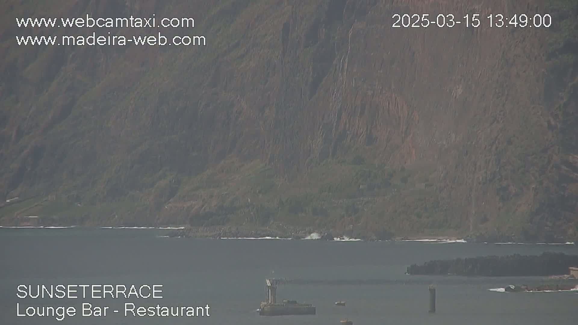 A hazy view shows a dark-colored rocky hillside sloping down to a calm body of water with a few small boats and a larger structure on the water.