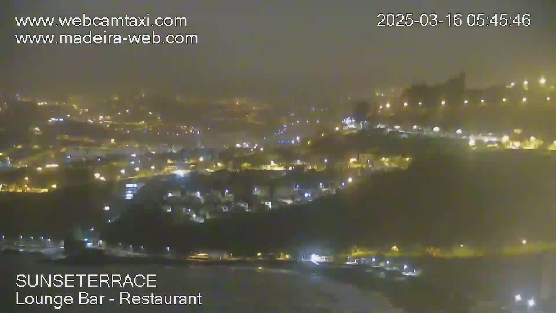 A nighttime view of a hazy, illuminated town situated on a hillside overlooking a dark body of water.