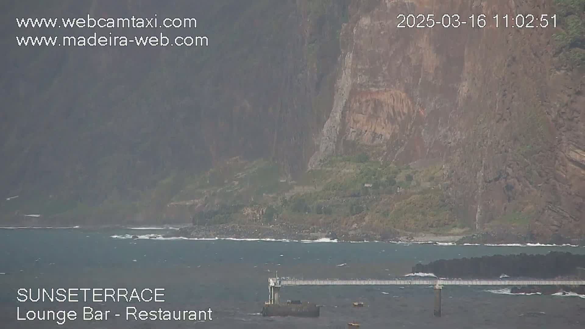 A long pier extends over a body of dark-grey water near a rocky, dark-brown cliffside on an overcast day.