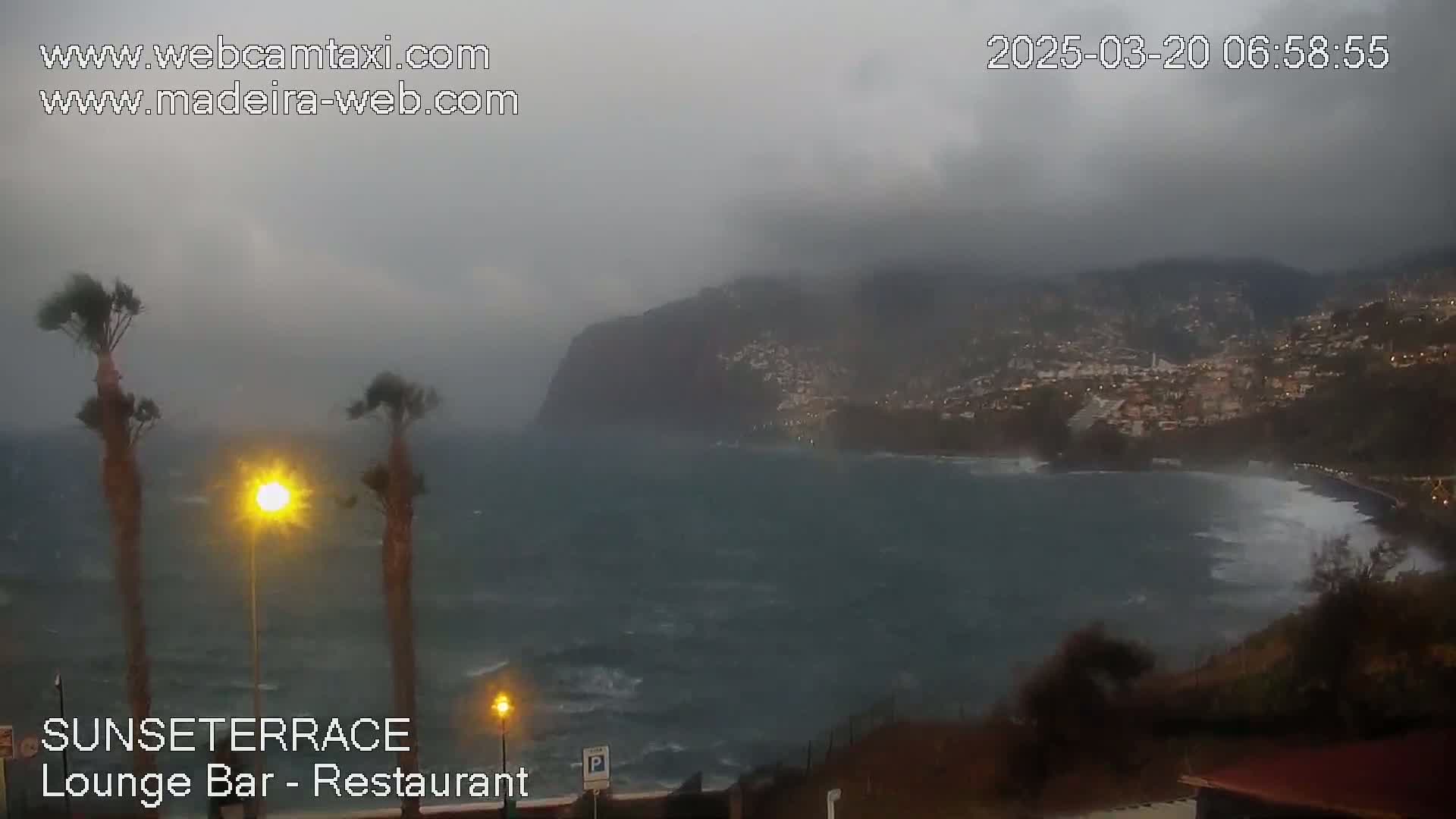 A stormy ocean scene shows a rocky cliffside with buildings, and palm trees in the foreground under an overcast sky.
