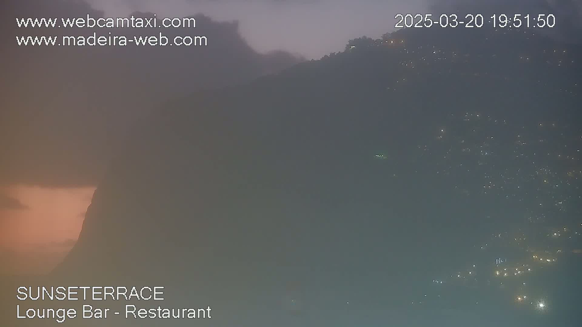 A hazy, dusky view shows a hillside at night, speckled with numerous small lights.