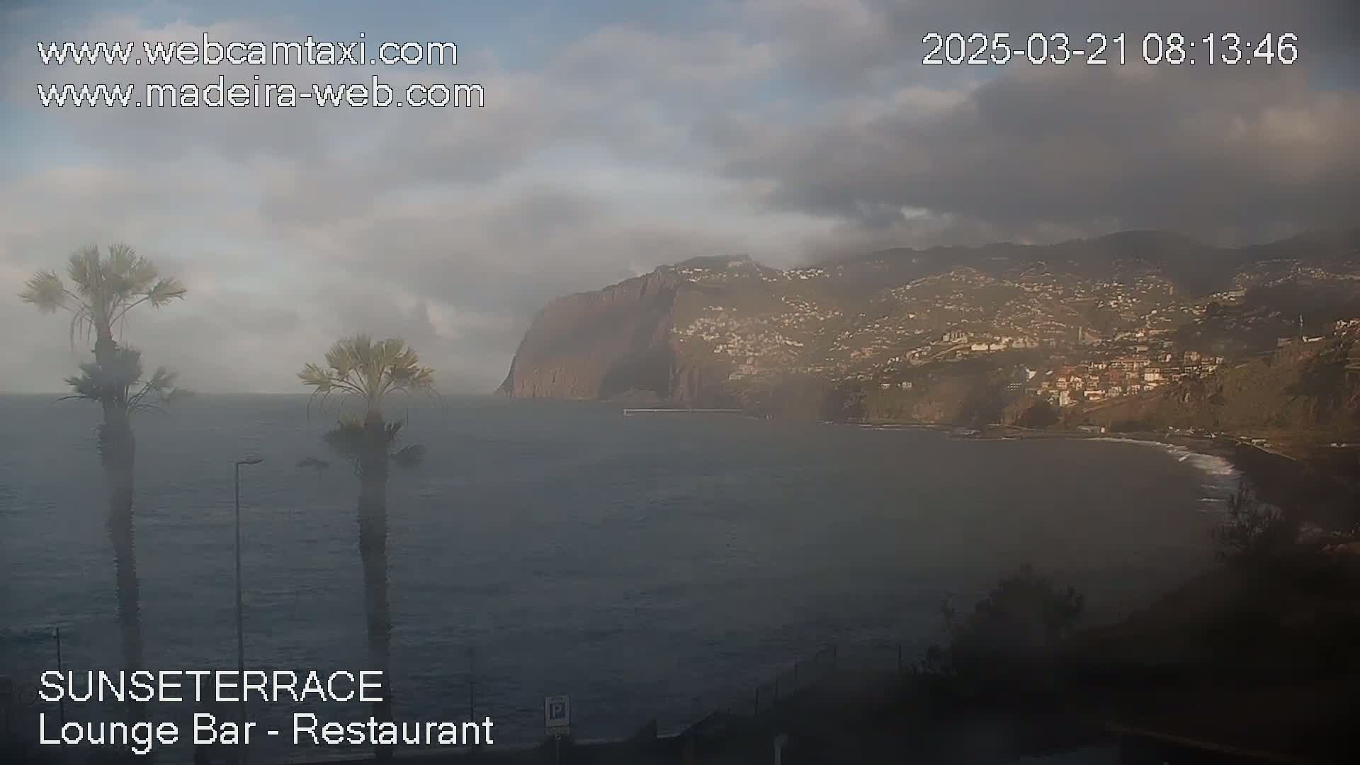 A hazy, overcast view of a calm ocean with a coastal town and a large cliff face in the background.