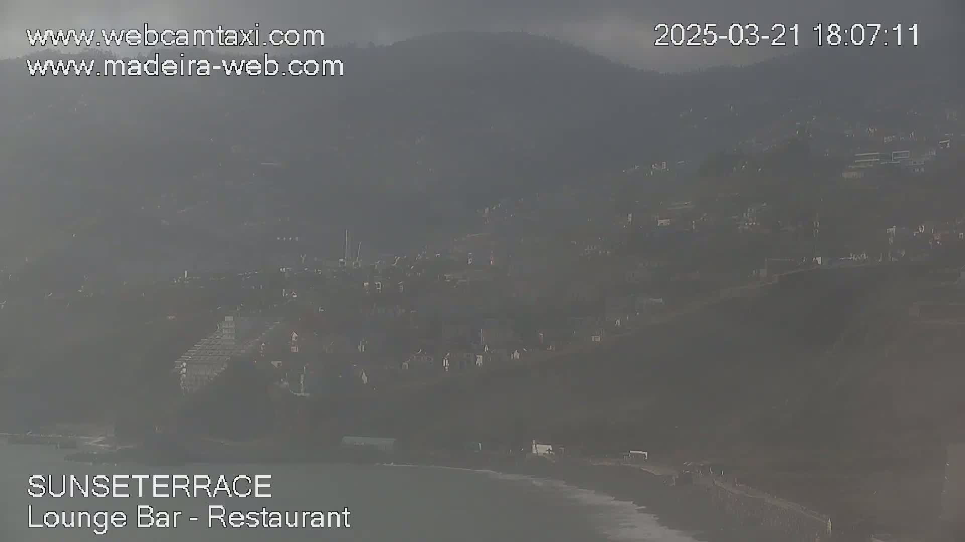 A hazy, overcast view shows a coastal town nestled on a hillside overlooking a dark ocean.