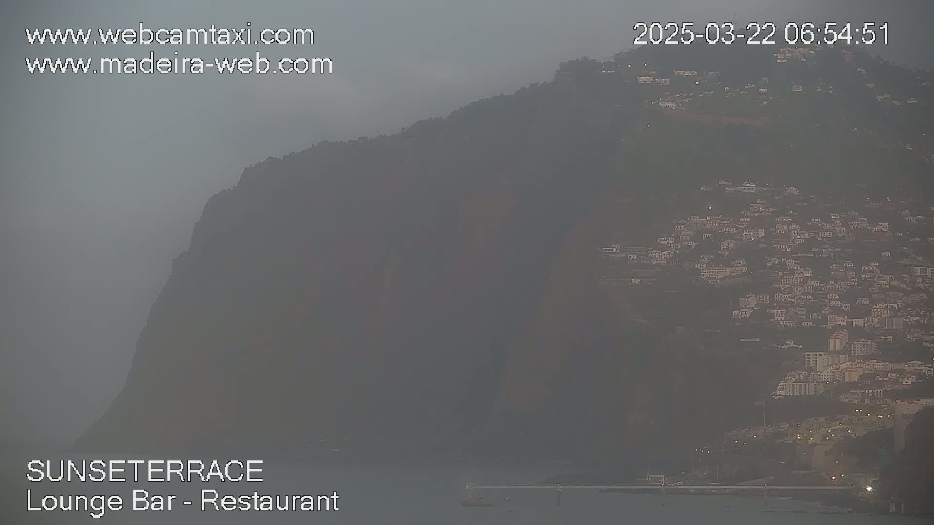 A hazy view shows a hillside town nestled against a large, dark mountainside.