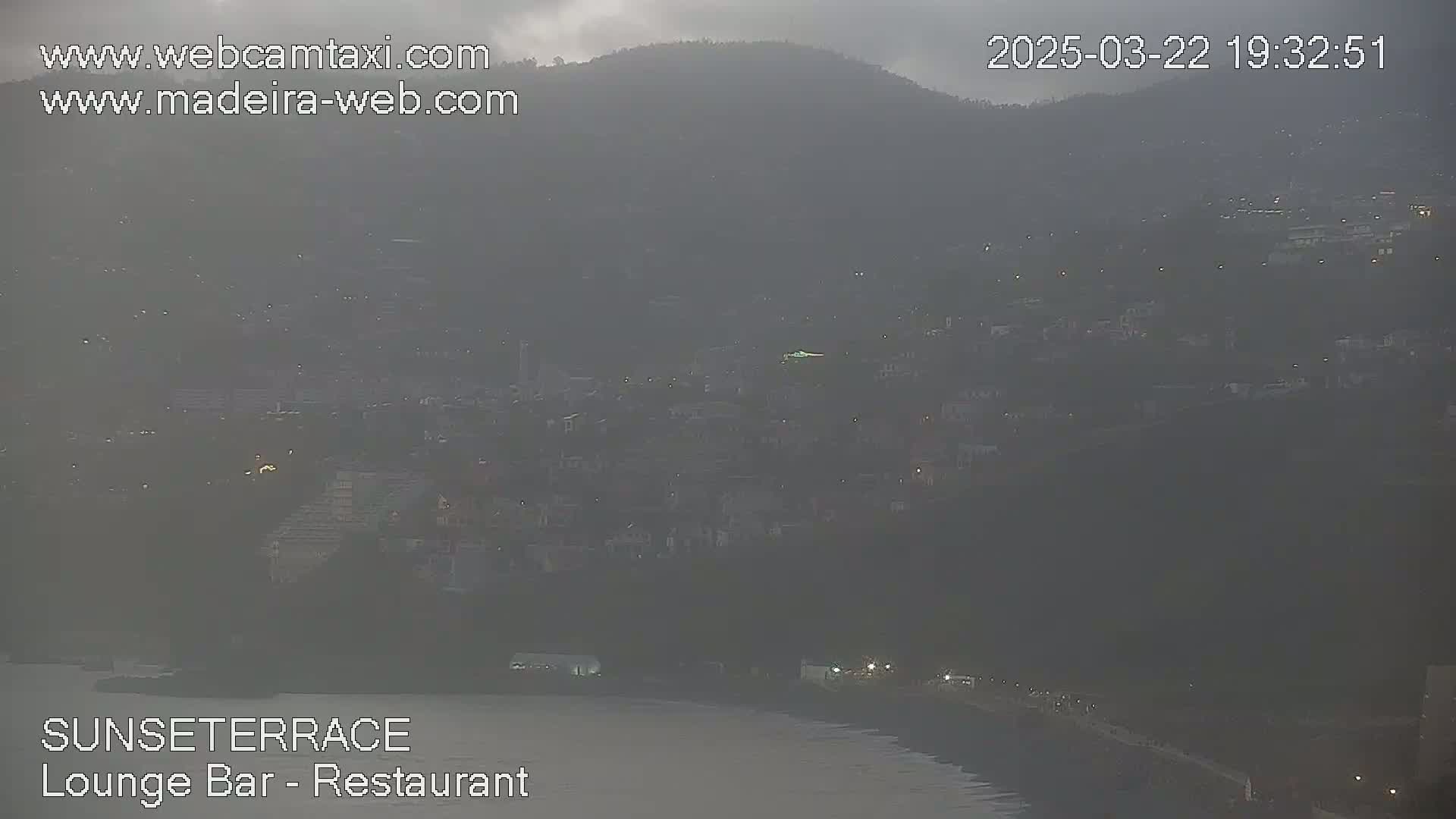 A hazy, dusk view shows a hillside town stretching down to a dark body of water.