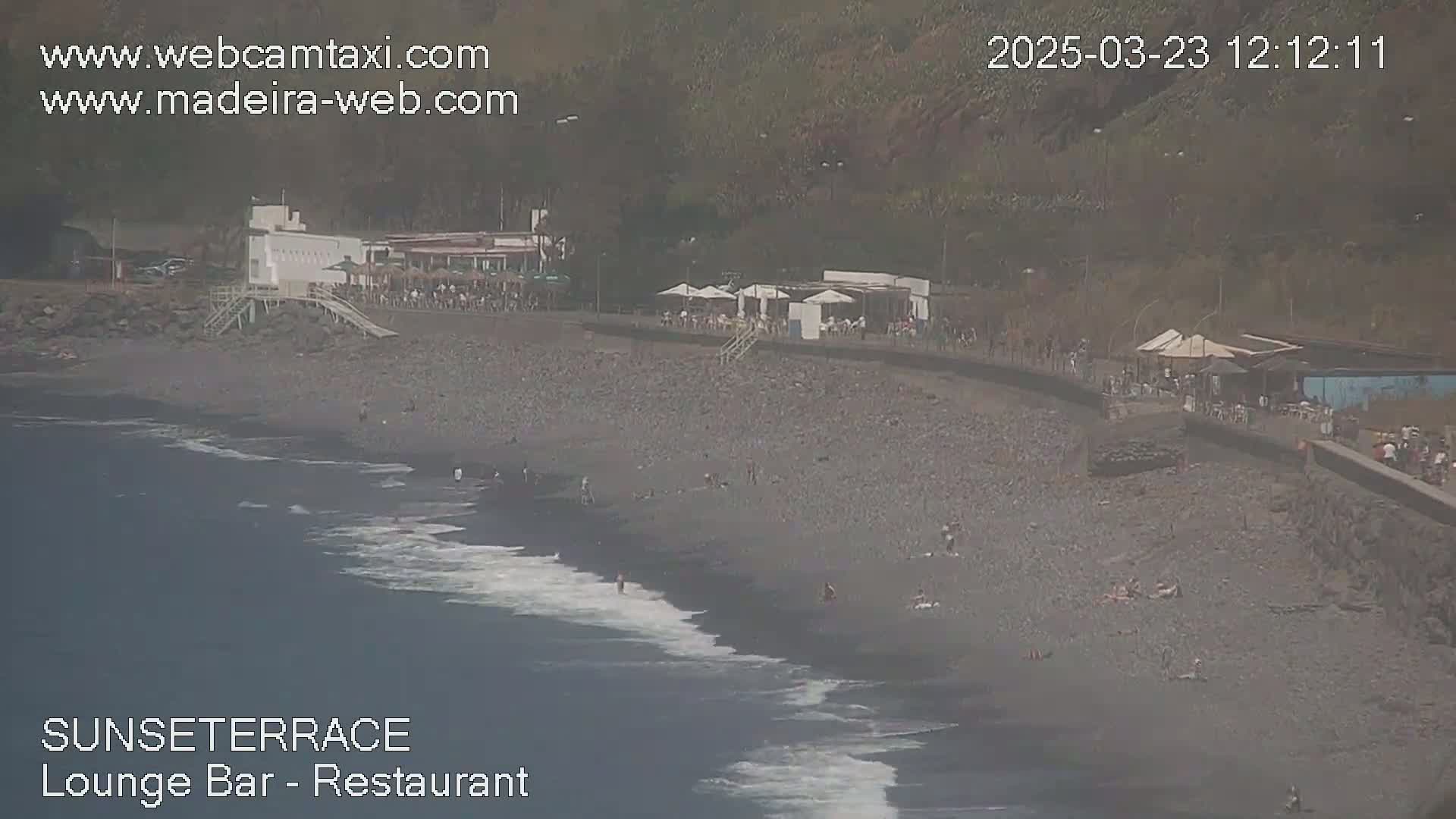 A dark-colored beach with people scattered along it and several buildings with outdoor seating situated above, all under a mostly clear sky.