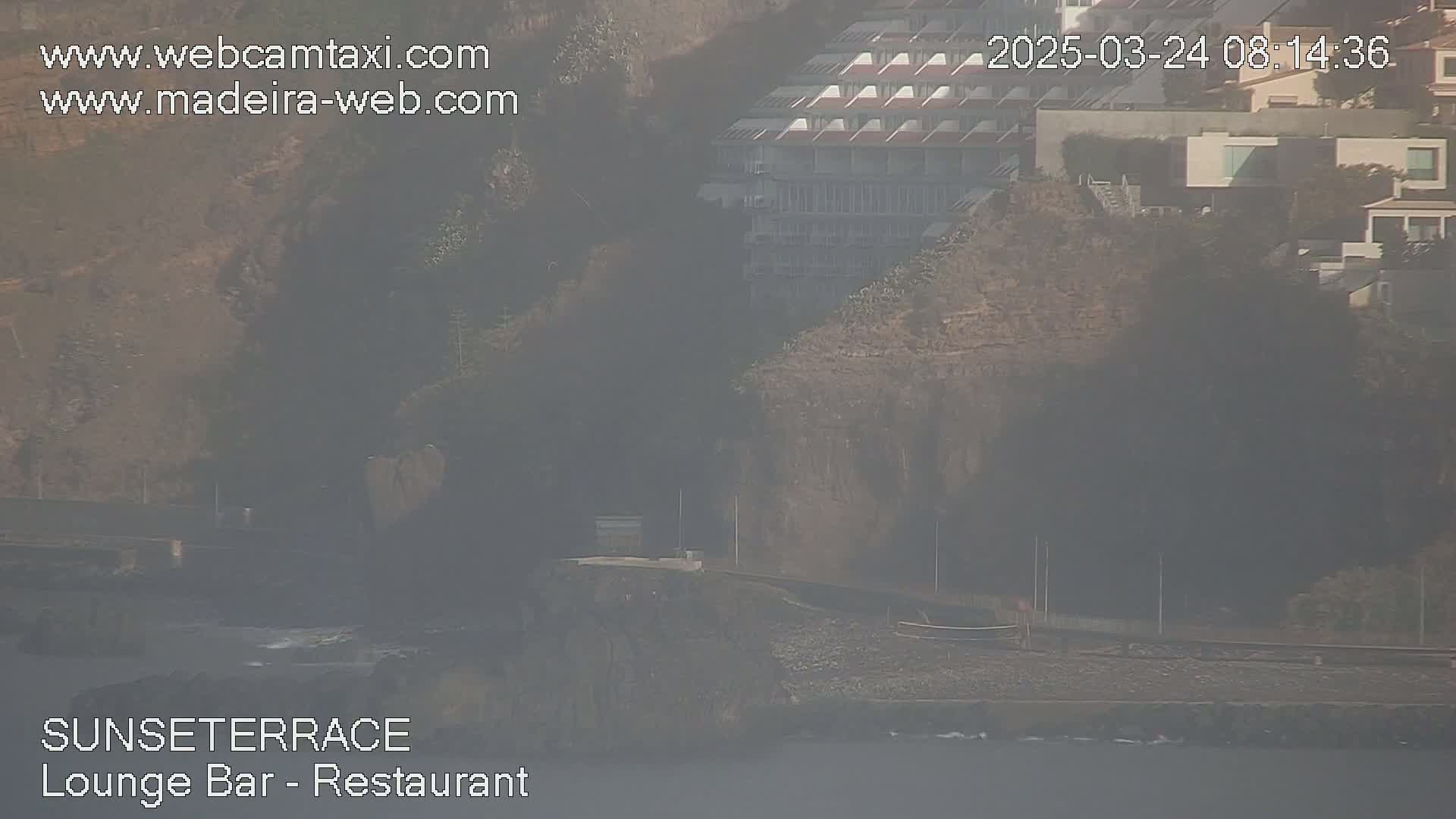 A hazy view shows a coastal area with buildings on a hillside overlooking a dark-colored beach and ocean.