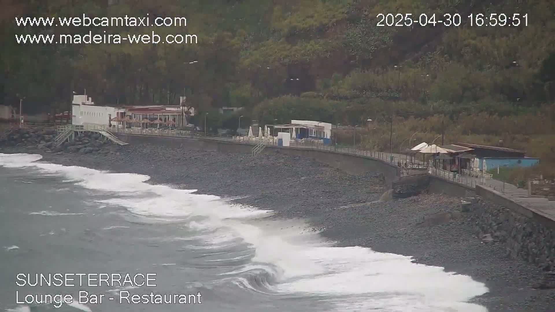 Ocean waves crash on a dark, pebbled shore next to a restaurant and walkway, backed by a hillside covered in dark green vegetation.