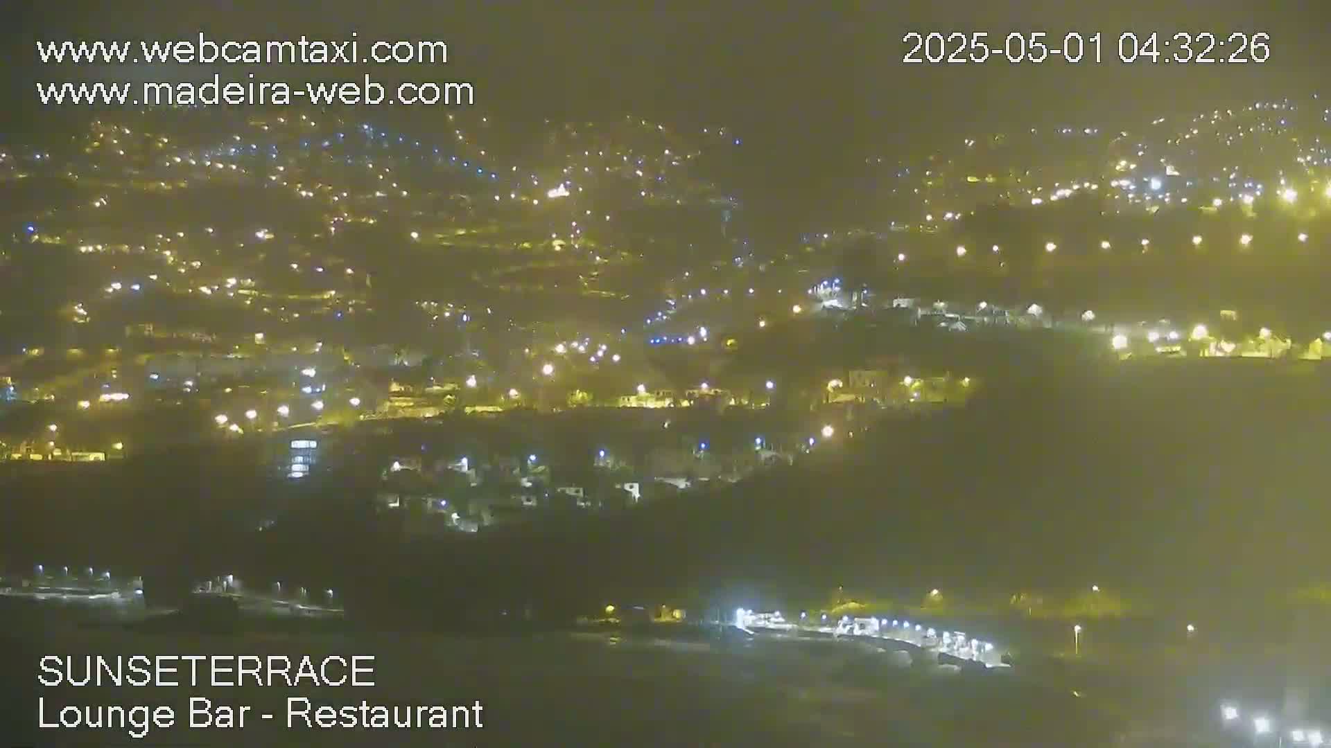 A hazy night scene shows a hillside town illuminated by numerous lights.