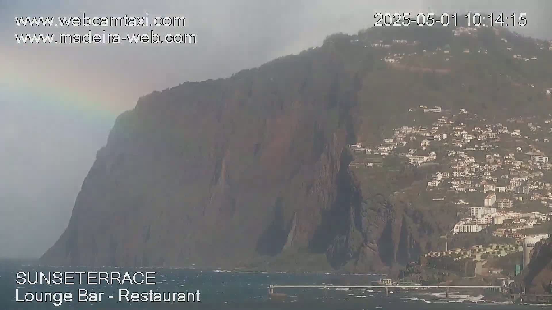 A hazy, partially obscured rainbow appears over a large, dark cliffside with a town nestled on its slopes overlooking a choppy ocean.