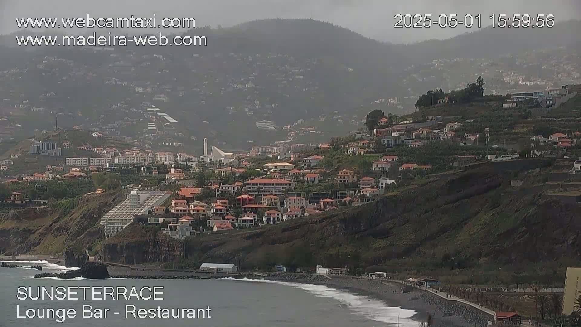 A hazy coastal town is situated on a steep hillside overlooking a dark-sand beach and ocean, under overcast skies.