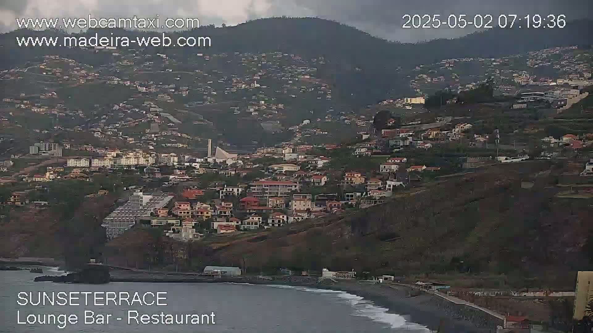 A hillside town overlooks a dark ocean under an overcast sky.