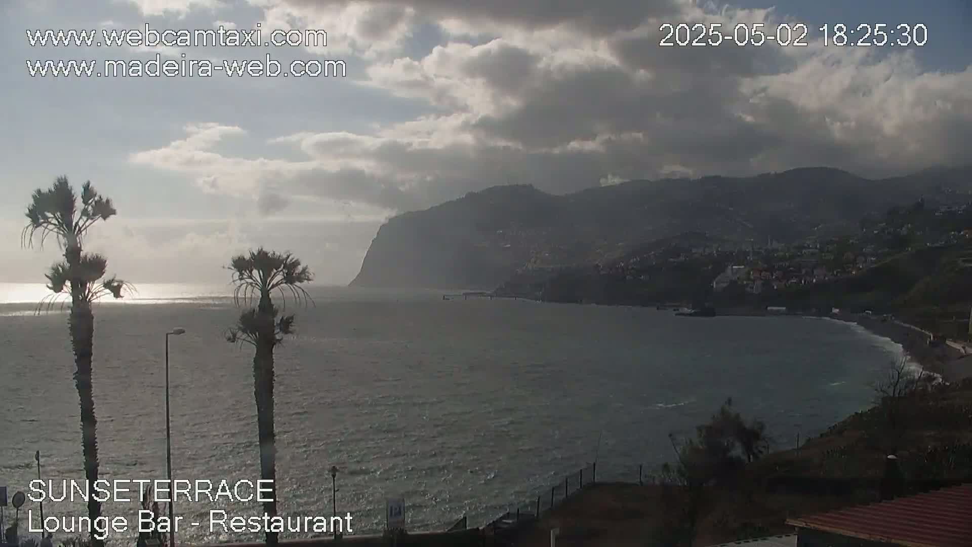 A partly cloudy day shows a coastal view of a dark-colored beach, calm ocean waters, and a mountainous region with a town built into the hillside.