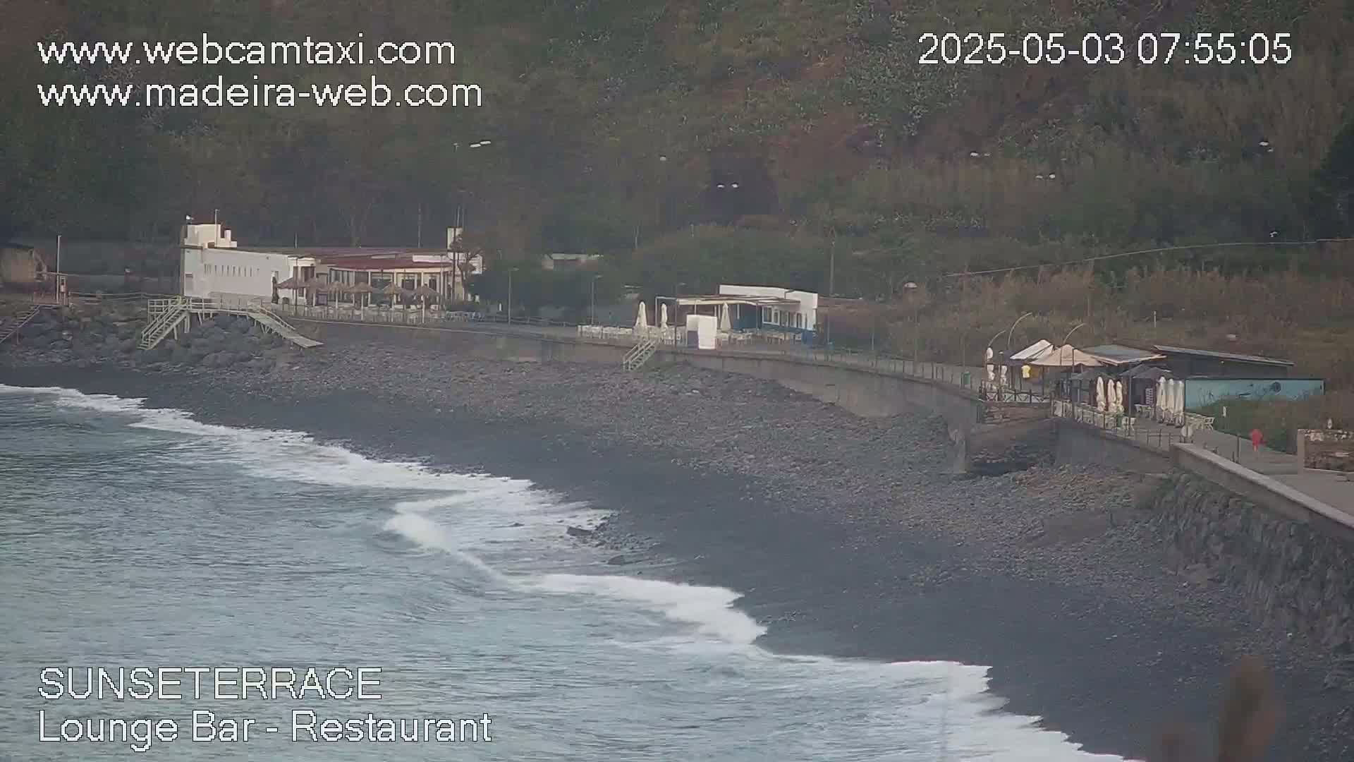 Ocean waves lap a dark, pebbled beach next to a building complex and a walkway, all set against a backdrop of a hillside.