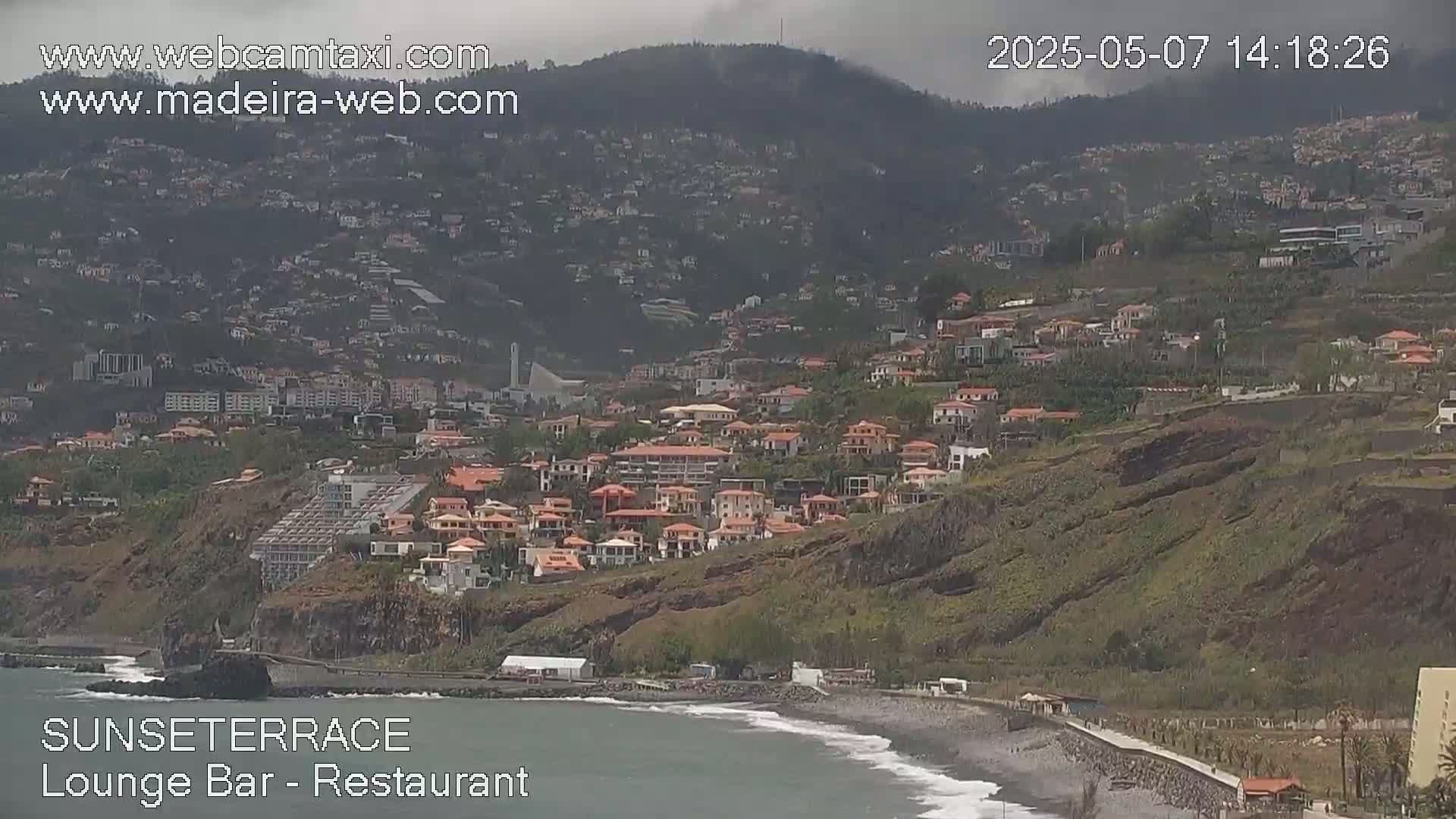 A coastal town built on a hillside overlooking a dark-sand beach and ocean under an overcast sky.