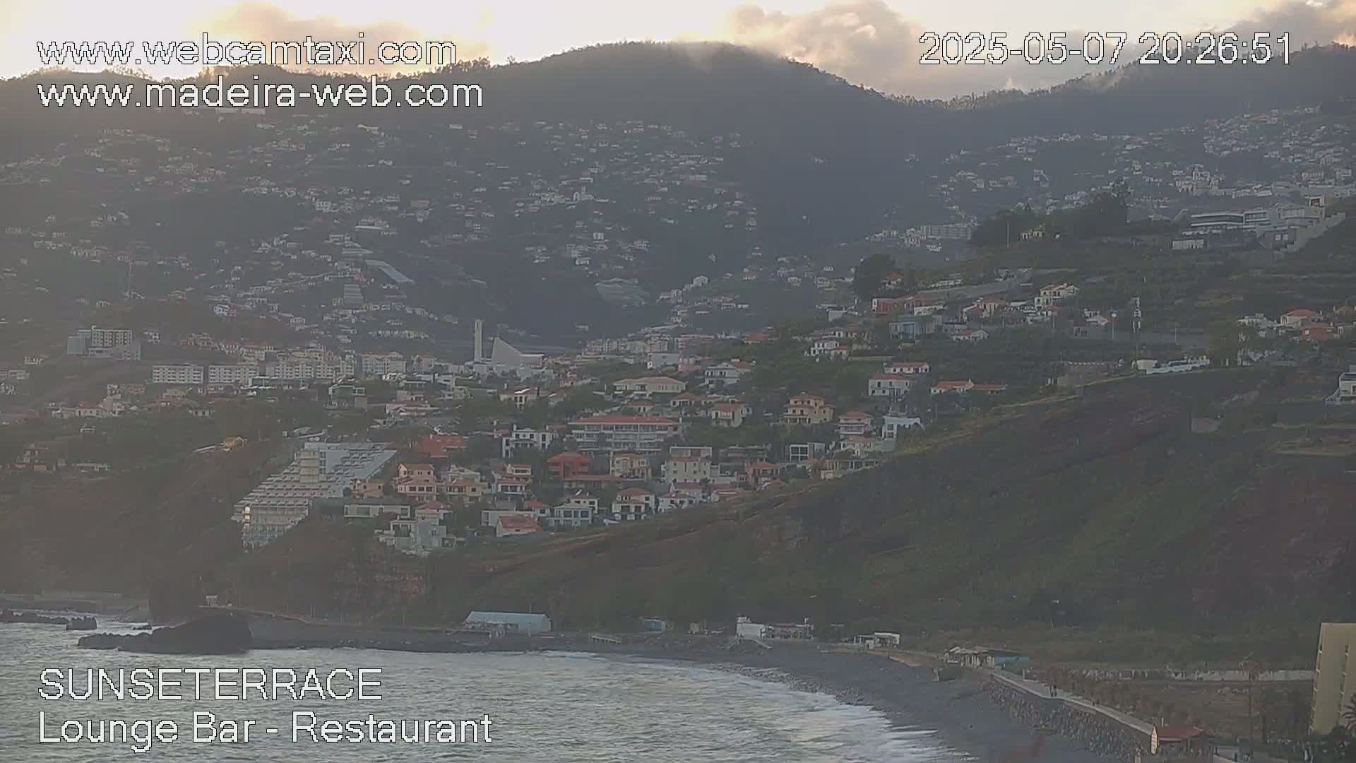 A hillside town overlooks a dark-sand beach and ocean, under a cloudy, dusky sky.