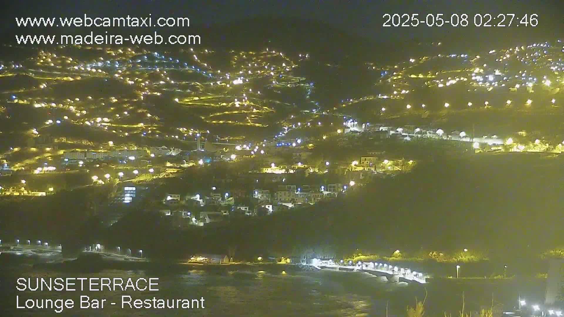 A nighttime view of a hillside town illuminated by many lights, overlooking a dark body of water.