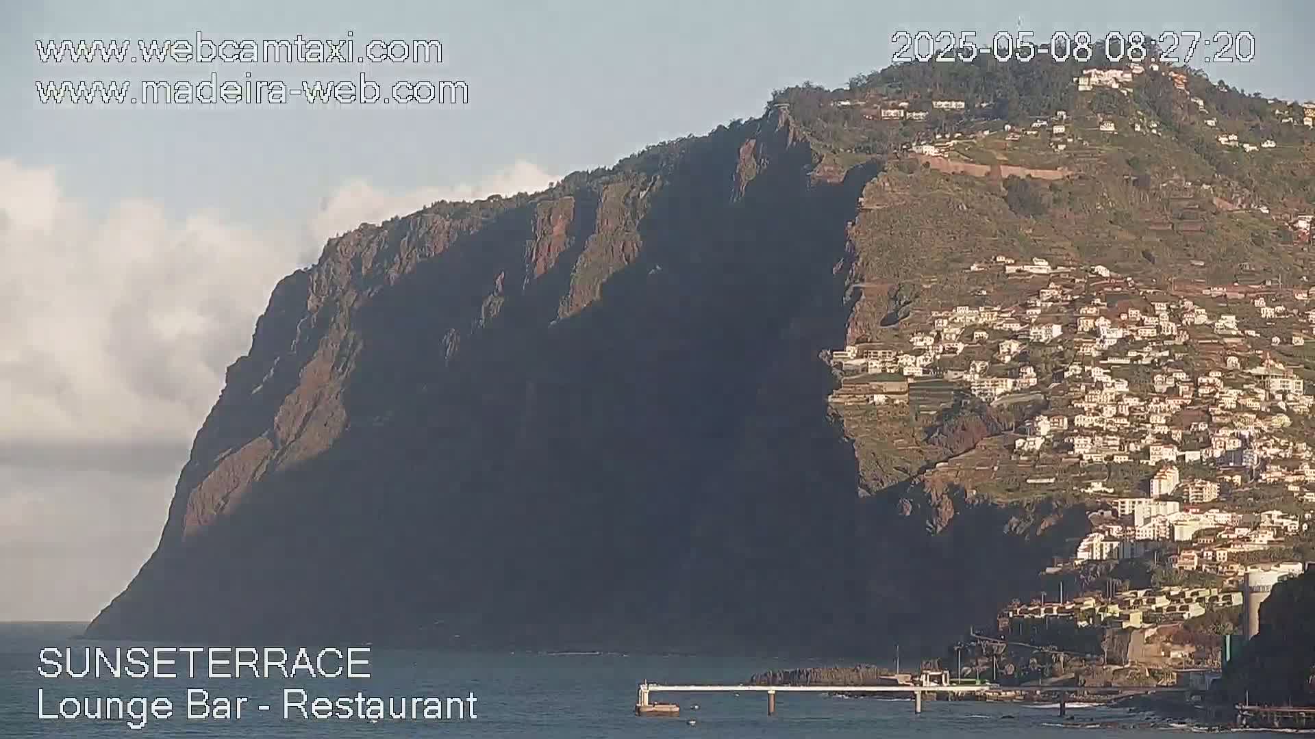 A coastal town clings to the side of a steep, dark cliff under a partly cloudy sky.