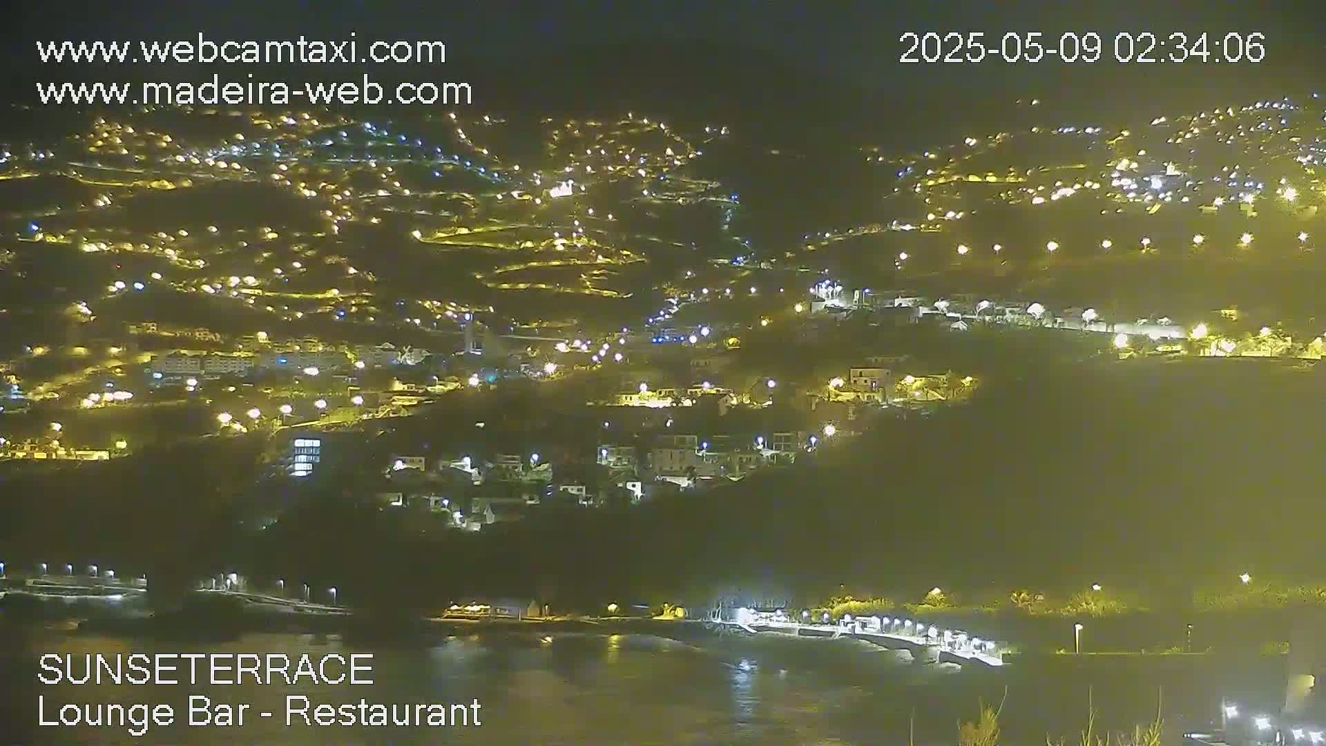 A nighttime view of a coastal town illuminated by many lights, overlooking a dark body of water.