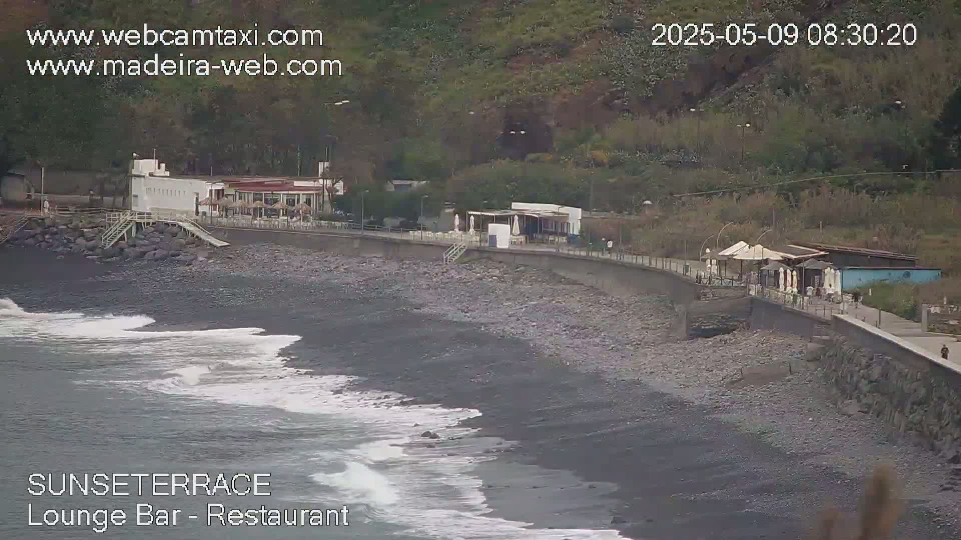A dark-sand beach with gentle waves laps against a seawall where buildings and outdoor seating areas are situated, backed by lush green hillsides.