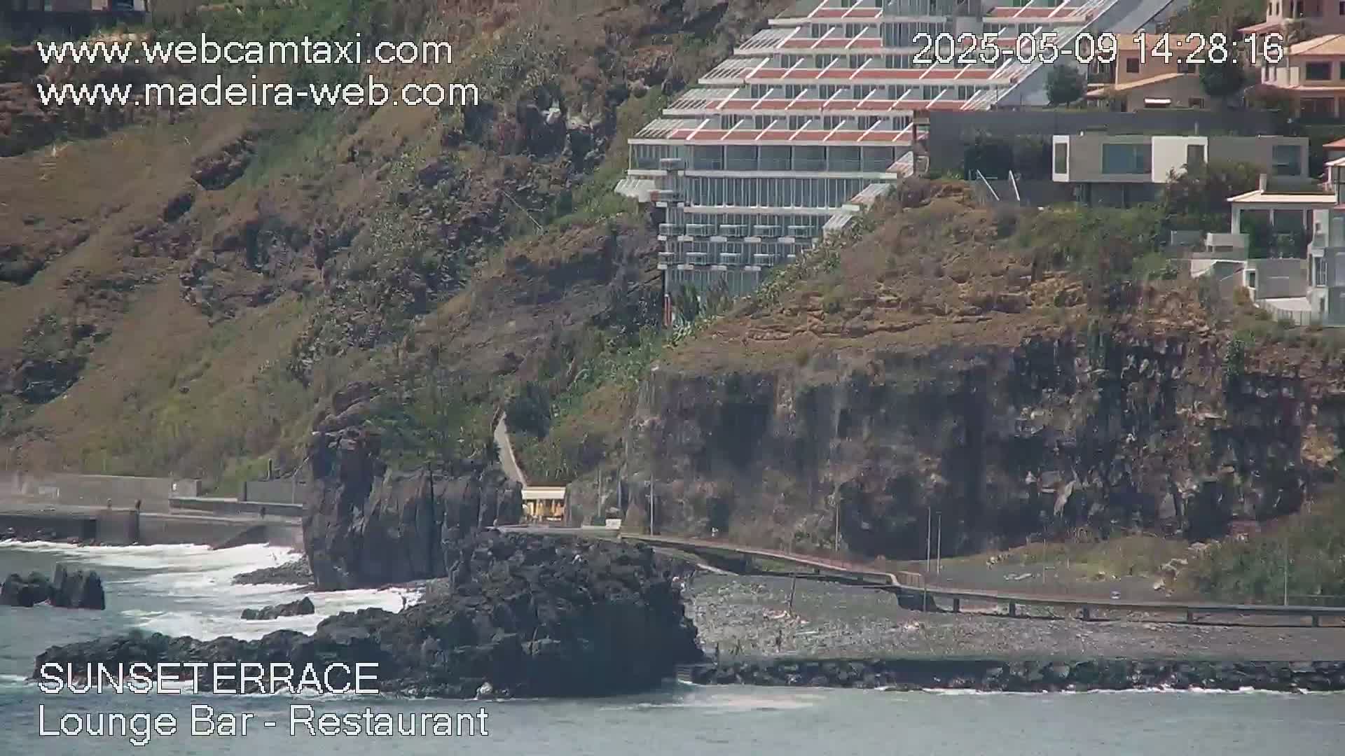 A seaside cliffside features a multi-story building, residential homes, and a rocky shoreline with a calm ocean.