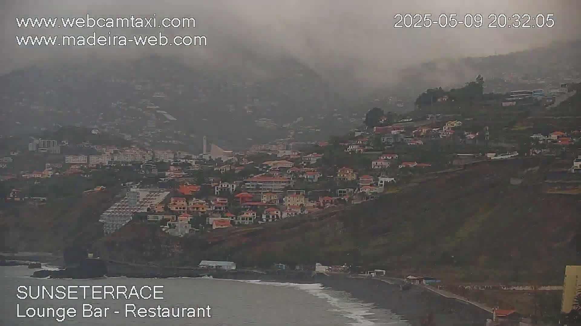 A coastal town nestled on a hillside is shrouded in mist and light rain, with a dark-colored beach visible at the bottom.