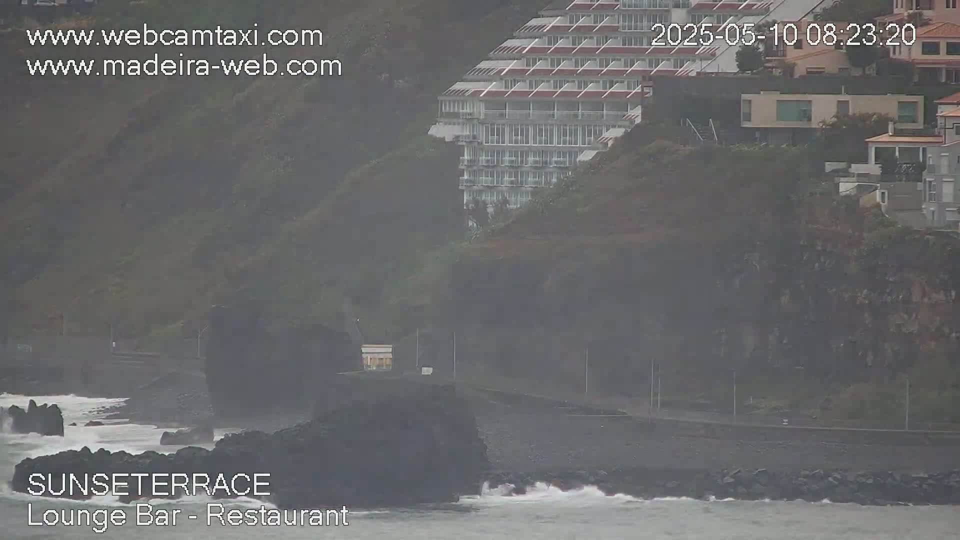 A multi-story building sits atop a cliff overlooking a rocky coastline with a hazy, overcast sky and moderate waves.