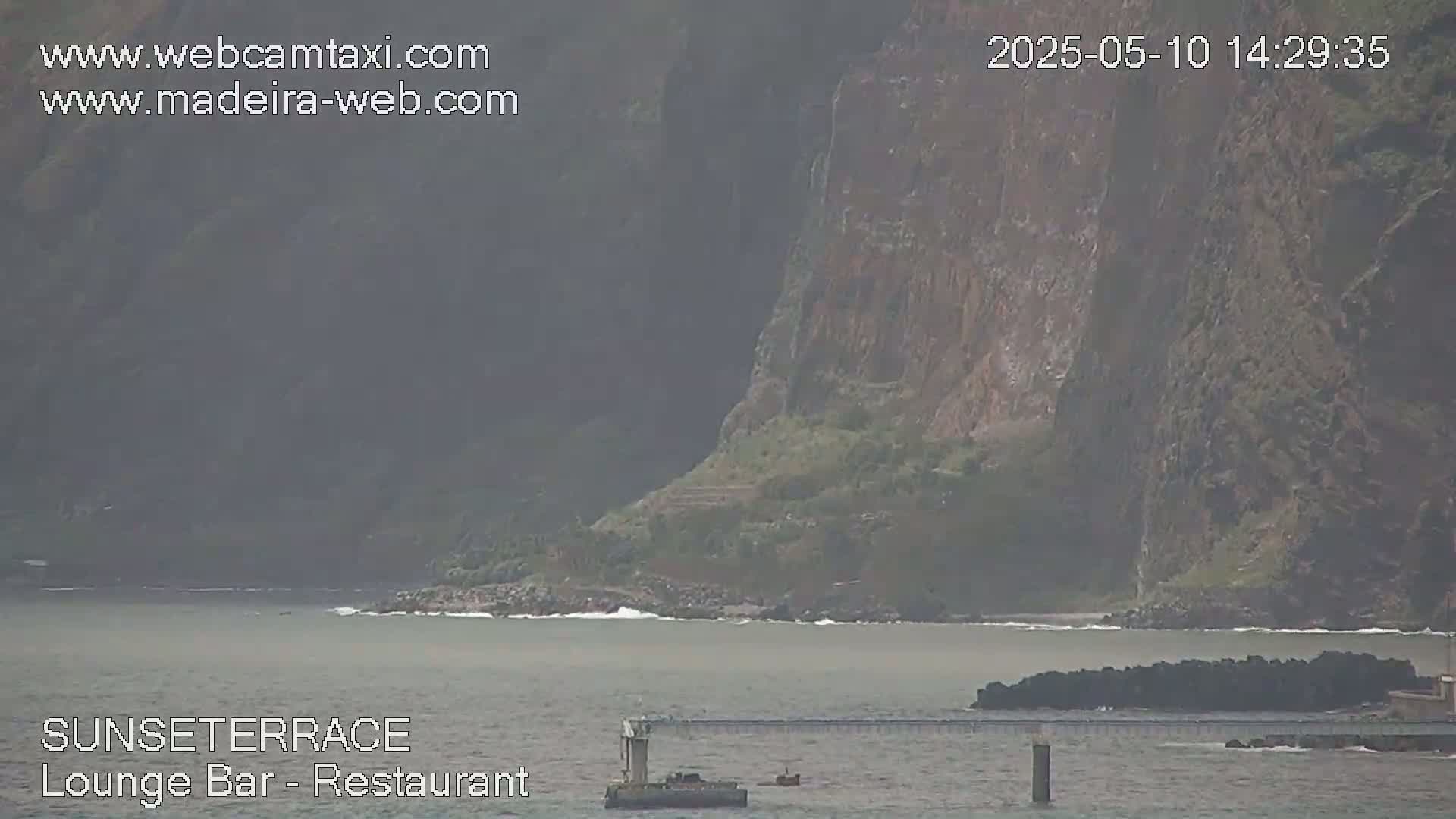 A hazy view shows a rocky cliff overlooking a calm body of water with a small island and a pier visible in the foreground.