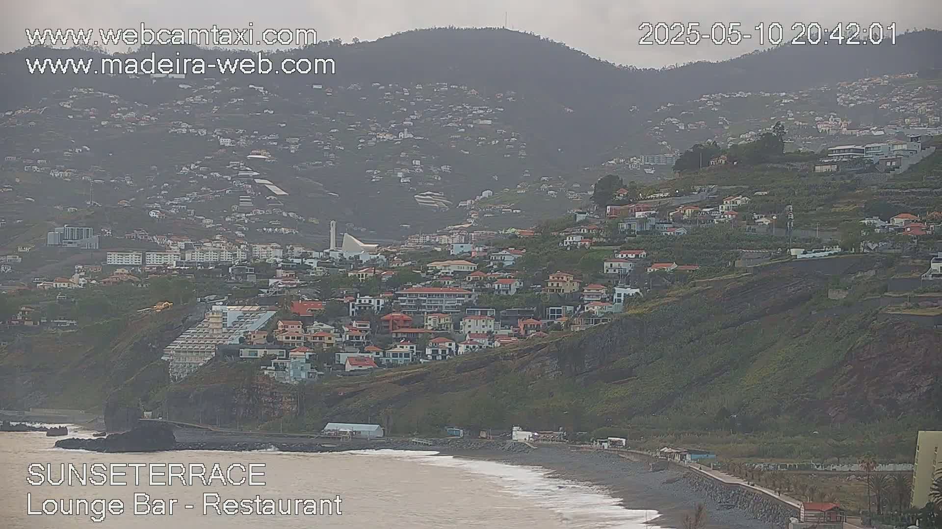 A coastal town built on a hillside overlooking a dark-sand beach with overcast skies.