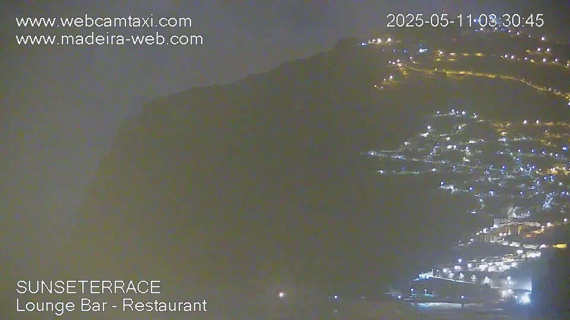 A nighttime long shot shows a hillside town illuminated by many lights under a dark, clear sky.