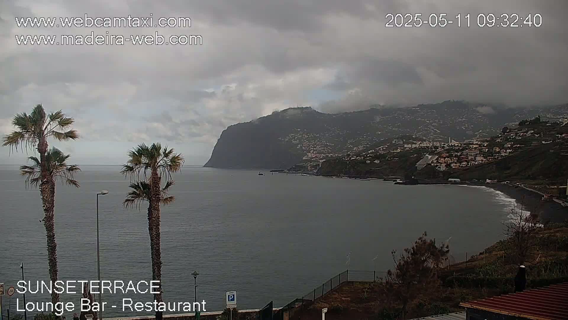 A coastal view on an overcast day shows a dark-colored body of water, a dark-colored beach, and a hillside town nestled beneath dark-colored mountains.