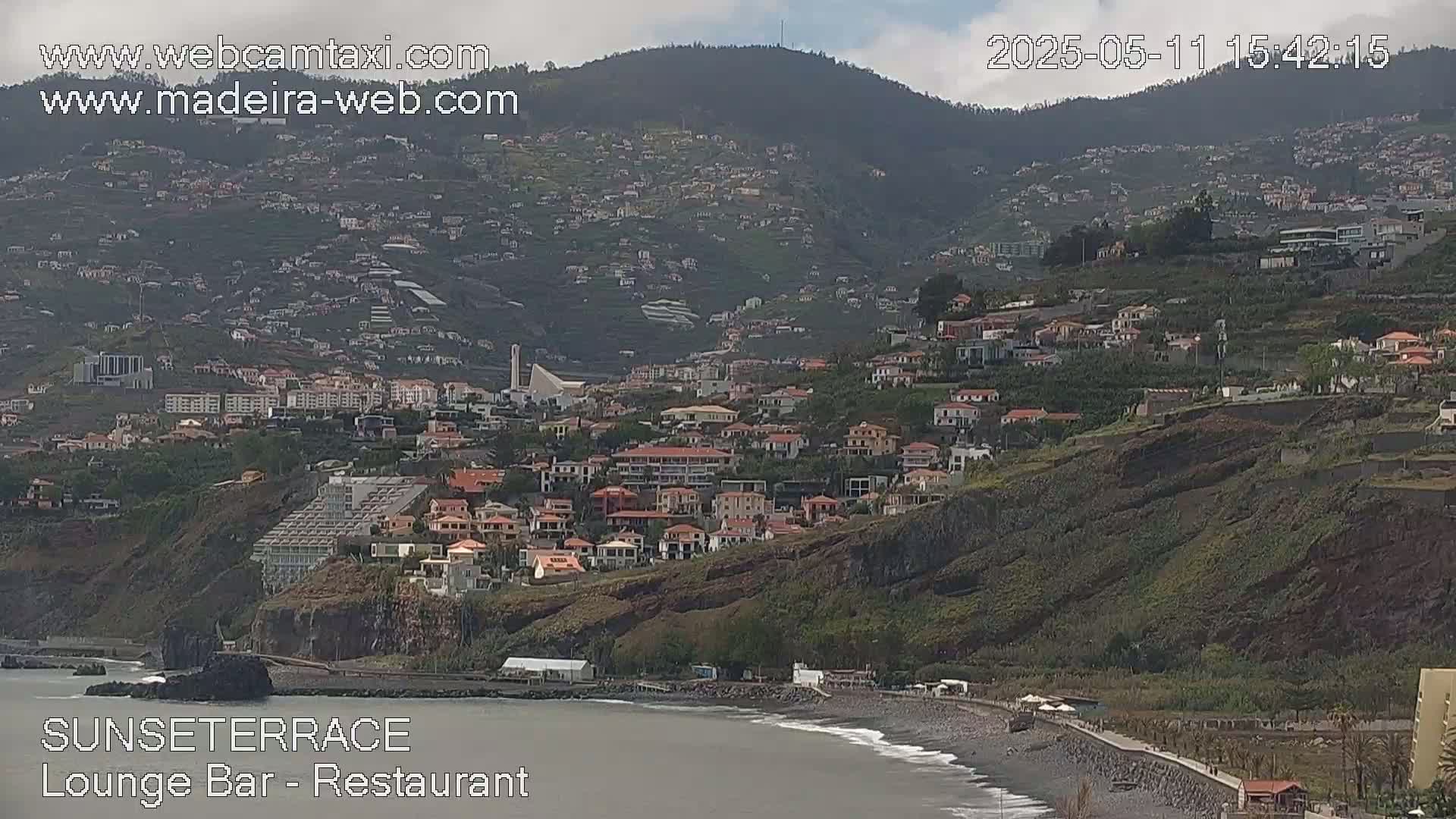 A coastal town nestled on a hillside below a mountain range under a partly cloudy sky, with a dark-sand beach at the water's edge.