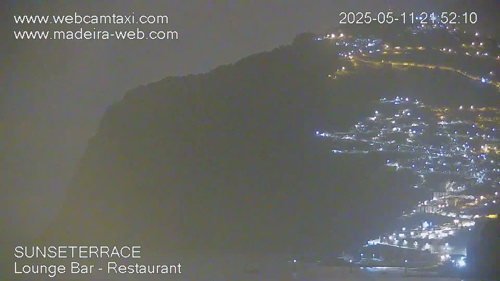 A nighttime, hazy view of a town nestled on a hillside, illuminated by many lights.