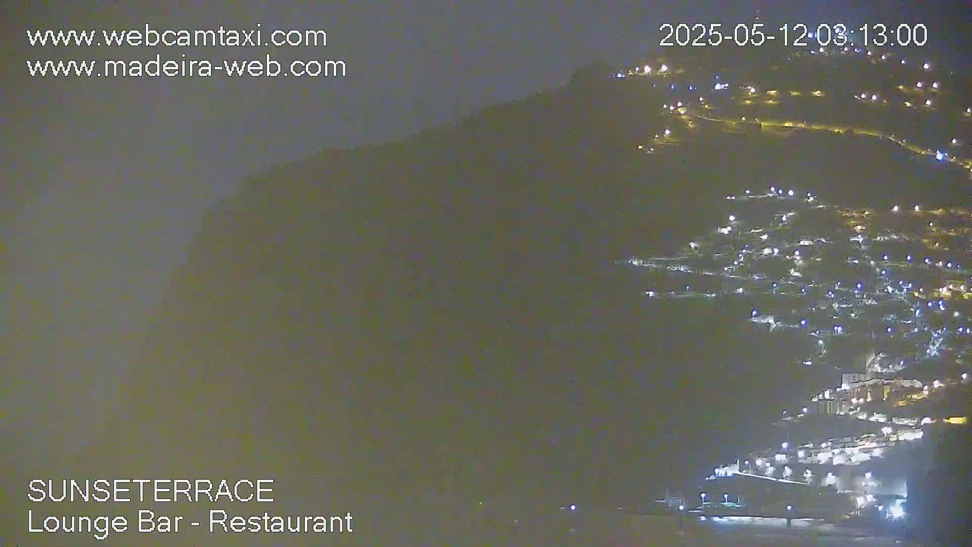 A nighttime long shot shows a hillside town illuminated by many small lights under a dark sky.