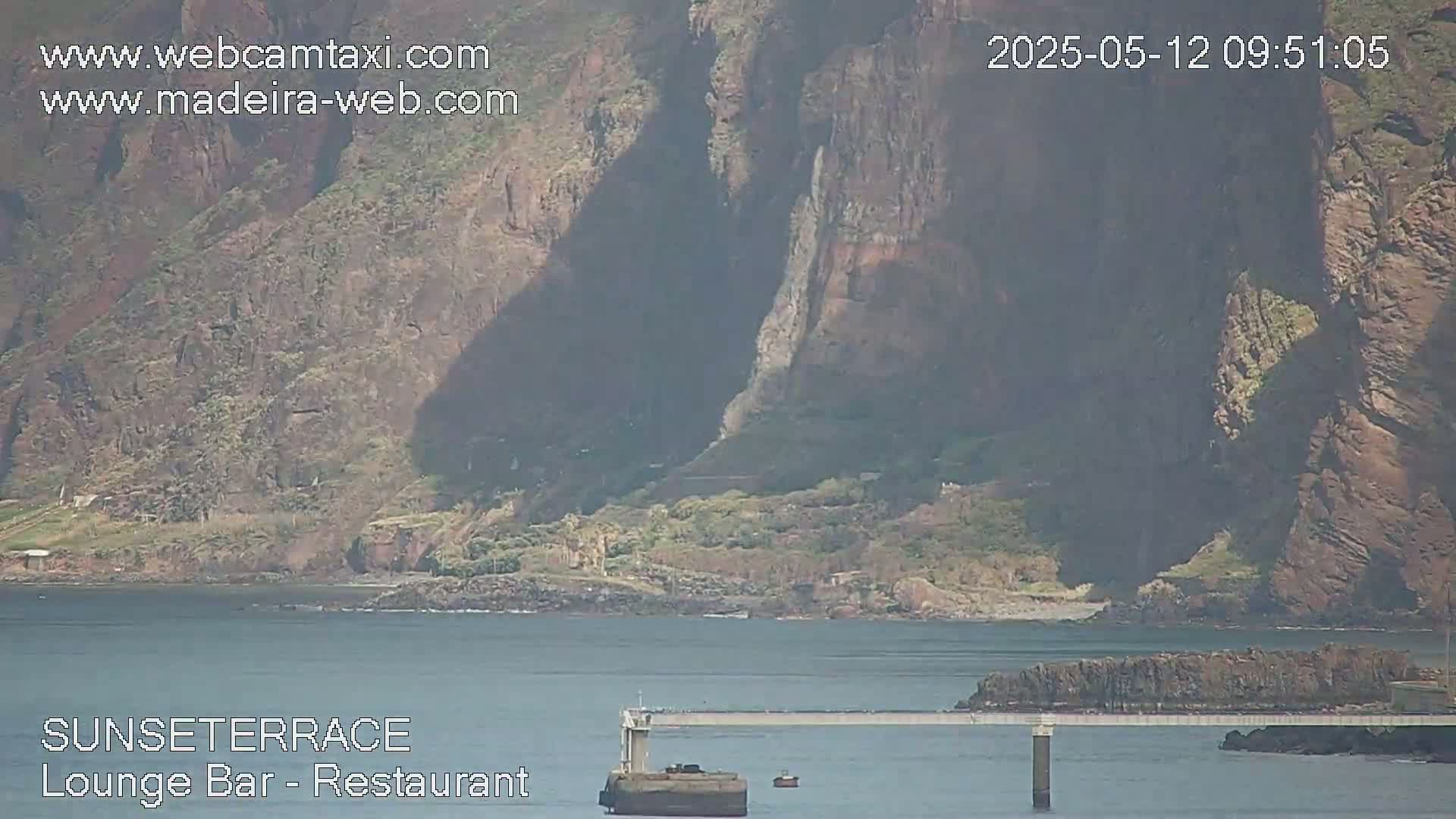 A long, low bridge extends from a rocky shoreline to a small island in front of a large, dark cliff face under sunny skies.