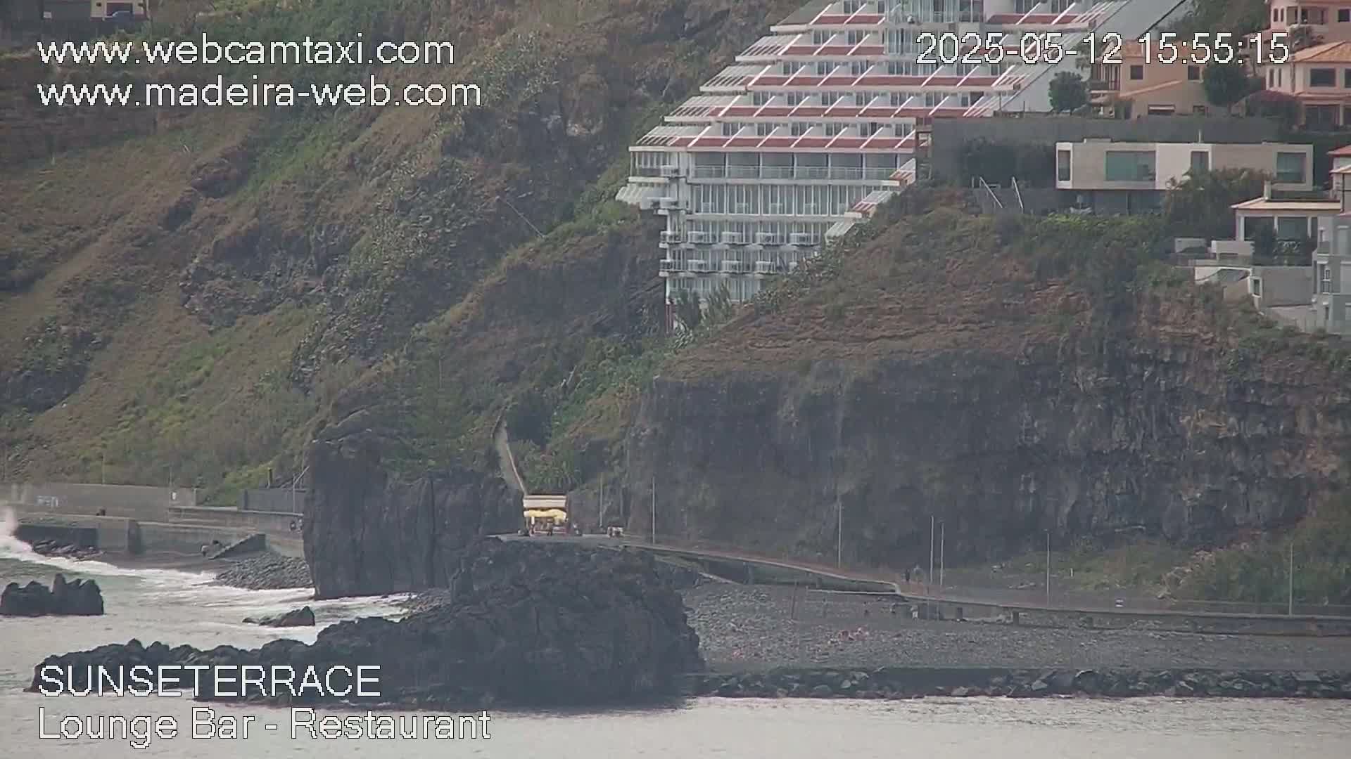 A coastal scene shows a large, multi-story hotel perched on a cliff overlooking a dark-sand beach with a rocky outcropping in the ocean; the weather appears overcast.