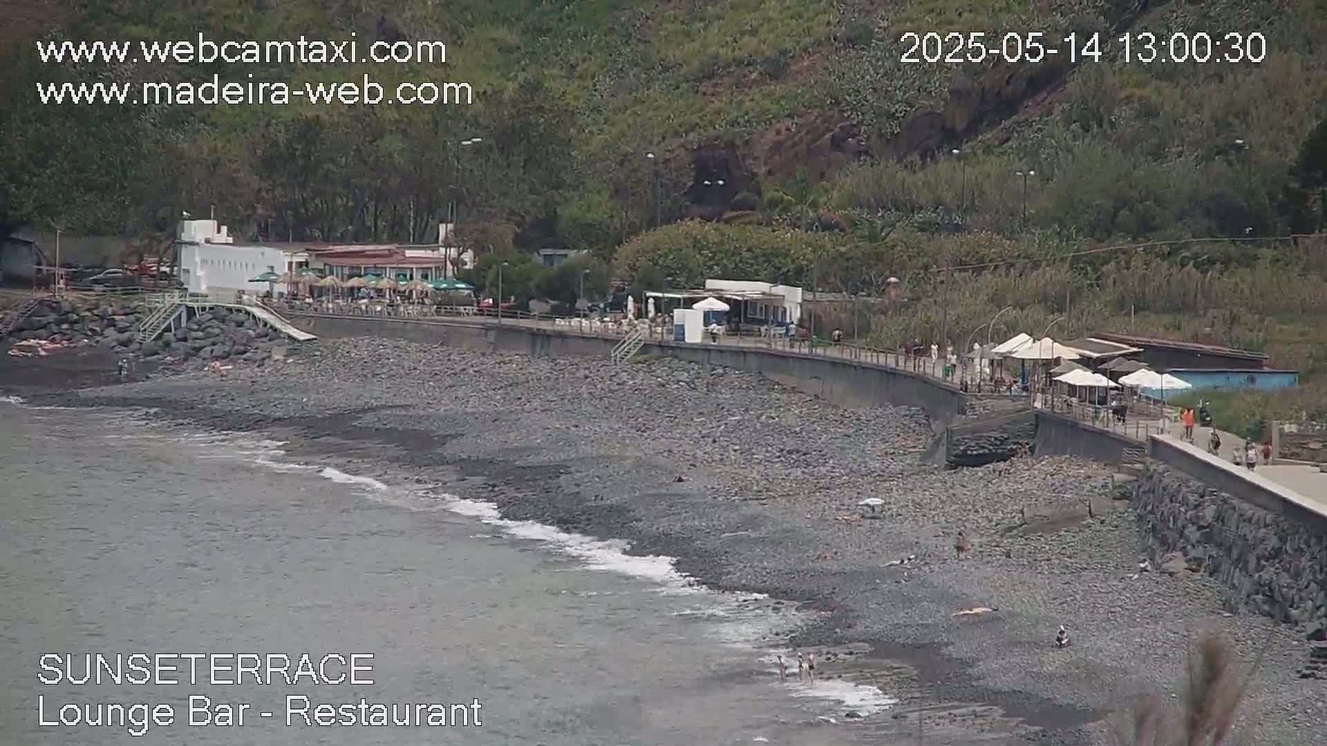 A rocky beach with a restaurant and bar sits below a green hillside on an overcast day.