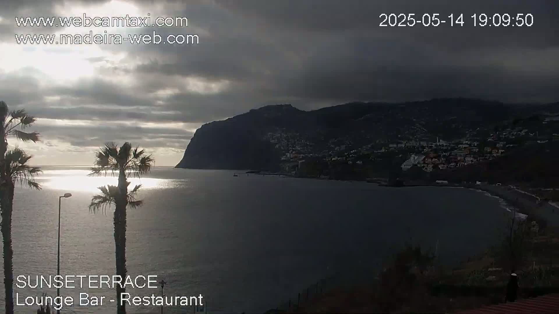 A dark, overcast sky looms over a calm ocean, with a large, dark mountain and a small coastal town in the background, and palm trees in the foreground.